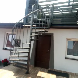 Zewnętrzne metalowe schody spiralne prowadzące na zadaszony taras, zamontowane przy białym budynku z brązowymi drzwiami i oknami.