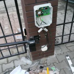 Montaż automatyki bramy przesuwnej: widoczny mechanizm FAAC przymocowany do słupka z cegły klinkierowej, otwarta obudowa elektroniki, przewody, narzędzia i instrukcja na brukowanej nawierzchni.
