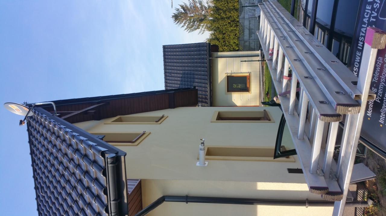 Antena satelitarna zamontowana na dachu domu jednorodzinnego z widoczną instalacją odgromową na ścianie, drabina oparta o samochód z reklamą usług instalacyjnych w tle.