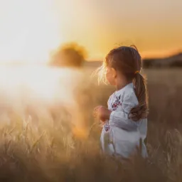 Dziewczynka w haftowanej koszuli, stojąca na polu zboża o zachodzie słońca, z włosami związanymi w kucyk.