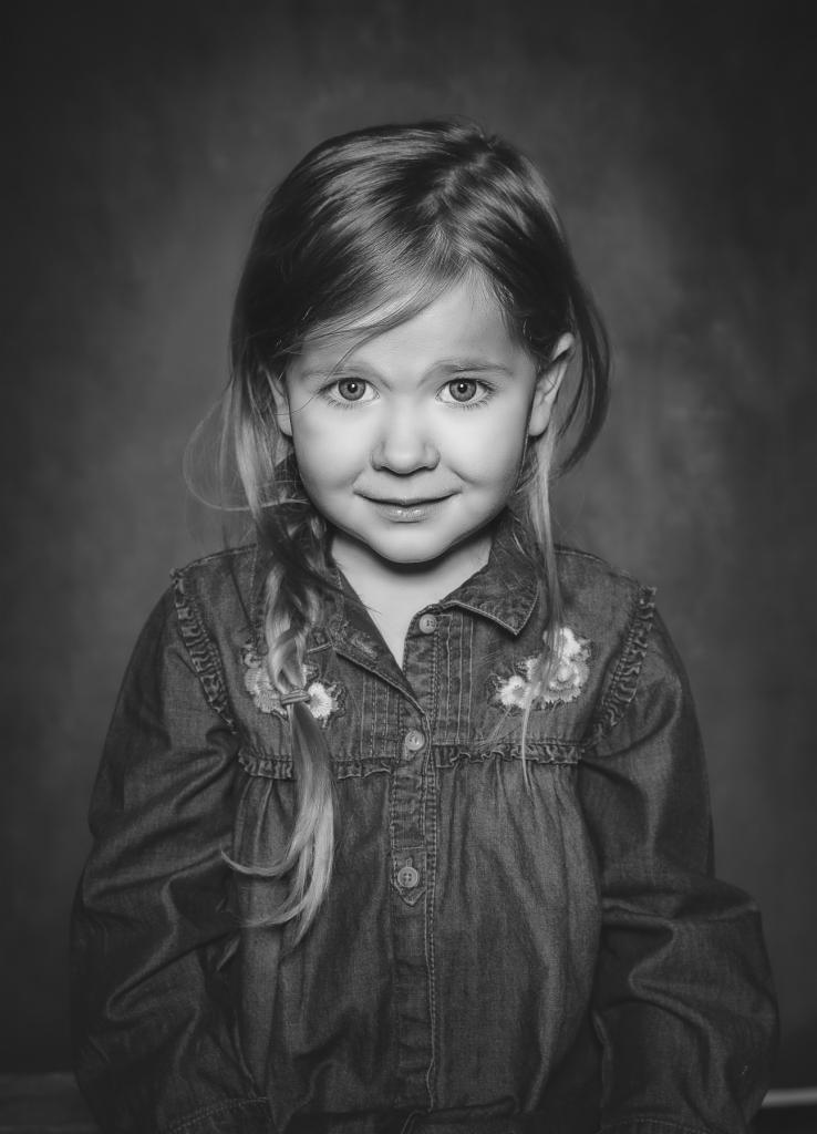 Portret uśmiechniętej dziewczynki w dżinsowej koszuli z kwiatowymi aplikacjami, włosy splecione w warkocz na tle ciemnego, jednolitego tła.