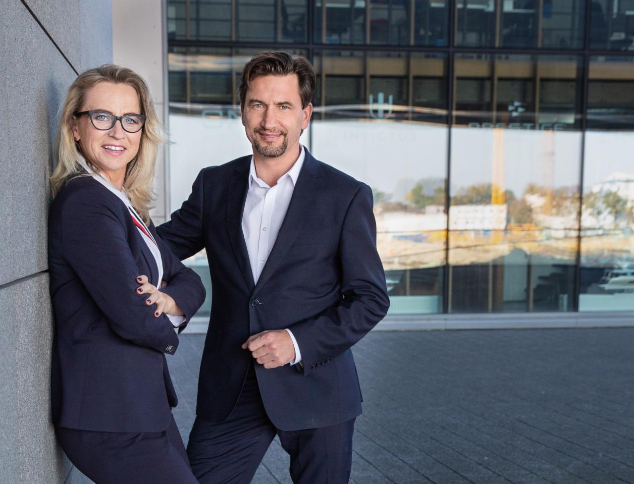 Portret biznesowy: kobieta w okularach oparta o ścianę, mężczyzna w garniturze z uśmiechem, nowoczesny budynek w tle.