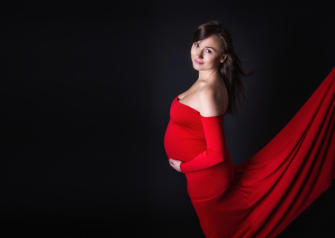 Kobieta w ciąży, ubrana w czerwoną suknię, pozuje na ciemnym tle, delikatnie uśmiechając się w kierunku obiektywu, z rękami ułożonymi na brzuchu.