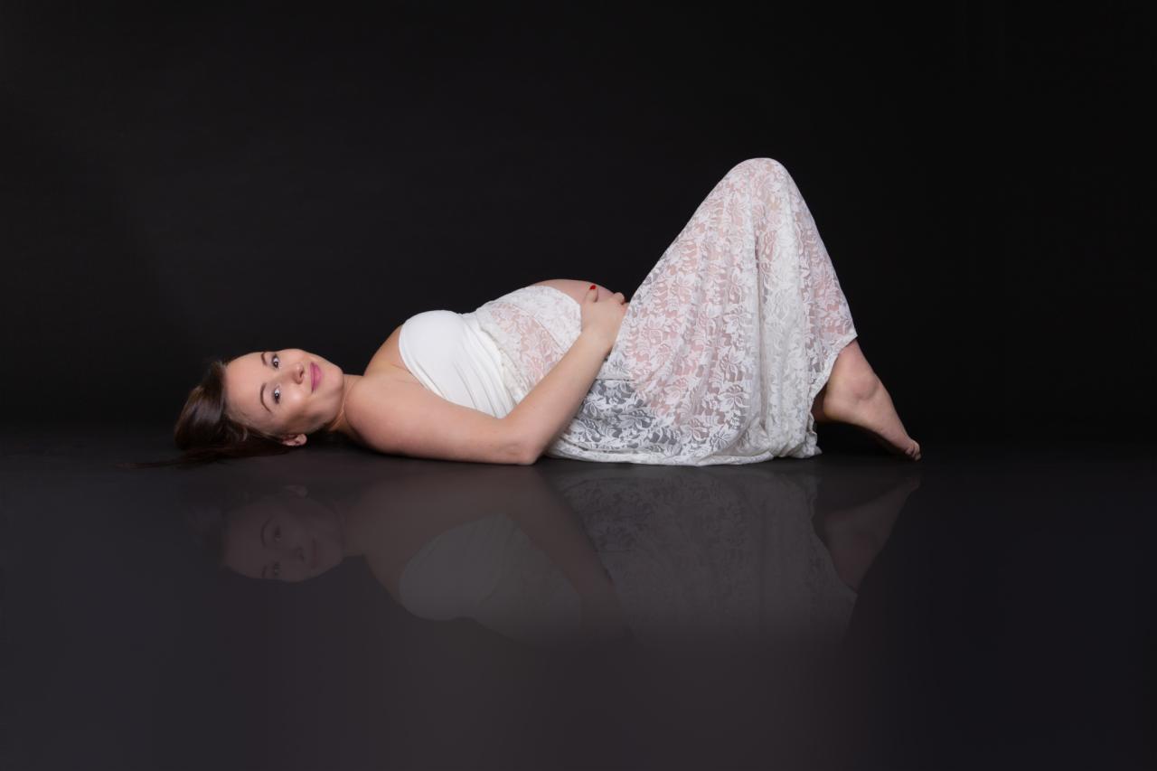 Kobieta w ciąży leży na boku w białej sukience z koronki na czarnym tle z odbiciem, trzymając ręce na brzuchu i uśmiechając się.