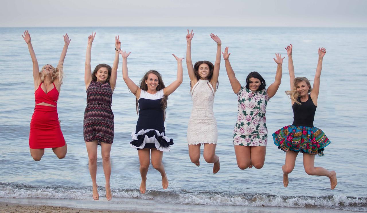 Sześć uśmiechniętych kobiet w sukienkach skacze radośnie na plaży, z morzem w tle.