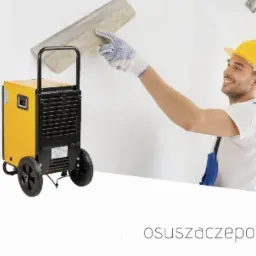 Dwa żółto-czarne osuszacze powietrza na kółkach, w tle dwóch mężczyzn w kaskach wyrównuje ścianę za pomocą pacy.