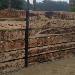 Drewniane ogrodzenie poziome z palonego drewna, wsparte na czarnych, metalowych słupkach, widoczne ślady po przypalaniu drewna, szara podmurówka.