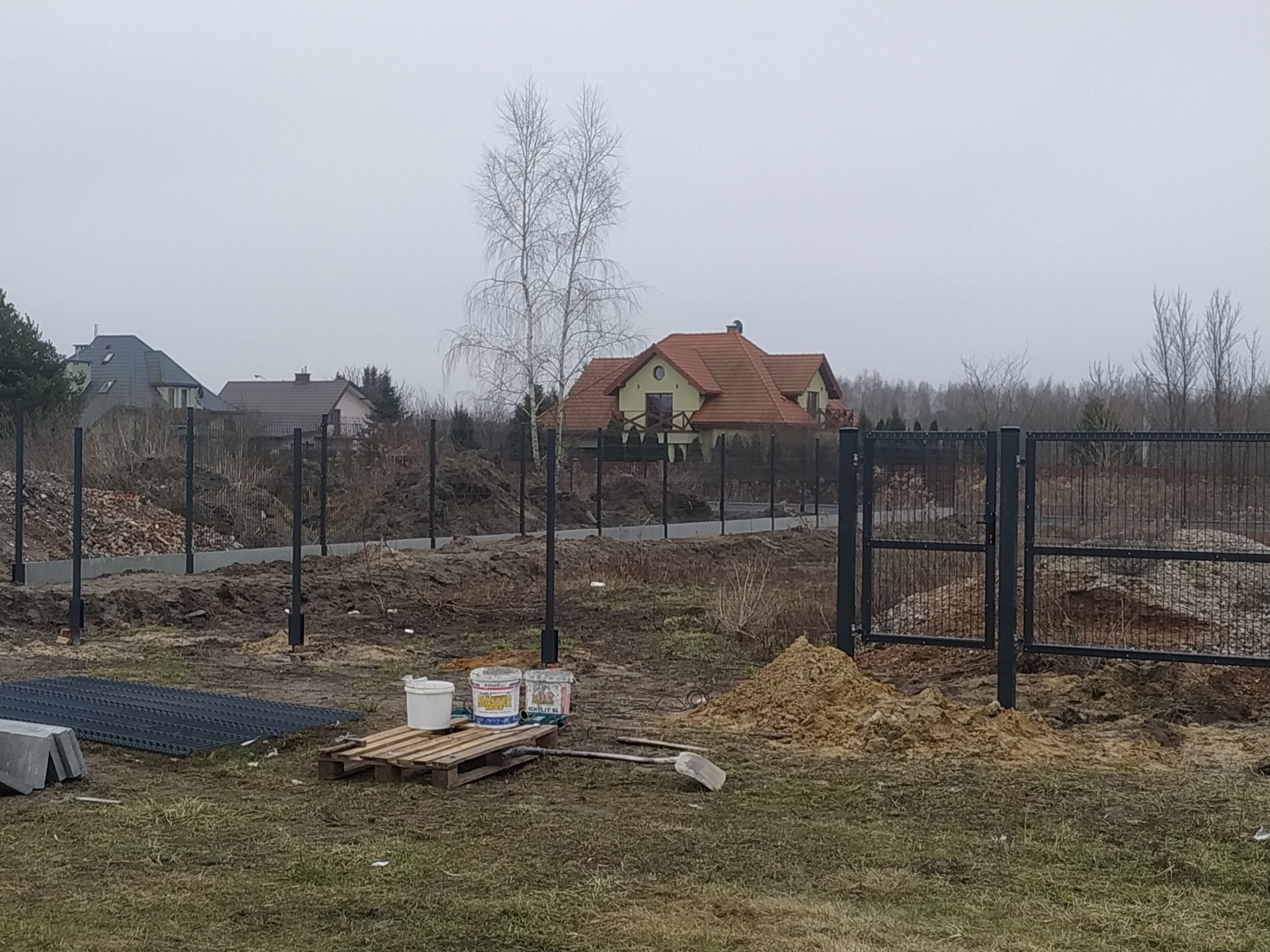 Etap budowy ogrodzenia panelowego z metalowymi słupkami na posesji, widoczne przygotowane panele, paleta z puszkami farby, łopata i pryzma piasku.