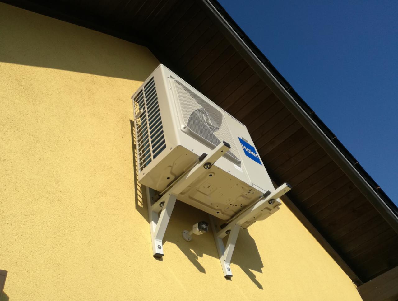 Zewnętrzna jednostka klimatyzacji Haier zamontowana na żółtej, teksturowanej ścianie budynku z widocznym fragmentem dachu i błękitnym niebem w tle.