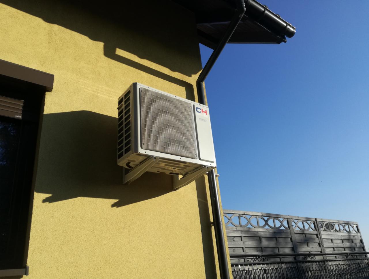 Zewnętrzna jednostka klimatyzatora zamontowana na żółtej ścianie domu, widoczne rynny i fragment ogrodzenia na tle błękitnego nieba.