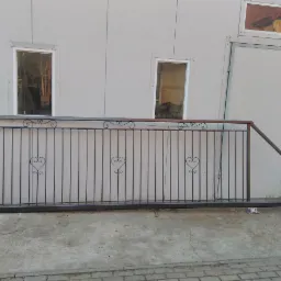 Fragment metalowej balustrady z dekoracyjnymi elementami w kształcie serc, ustawionej przed budynkiem o jasnej elewacji z widocznymi oknami i otwartymi drzwiami.