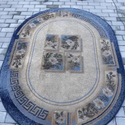 Owalny dywan z niebieskimi i beżowymi wzorami, leżący na szarej kostce brukowej, obok odkurzacza piorącego Karcher z końcówką do czyszczenia dywanów.