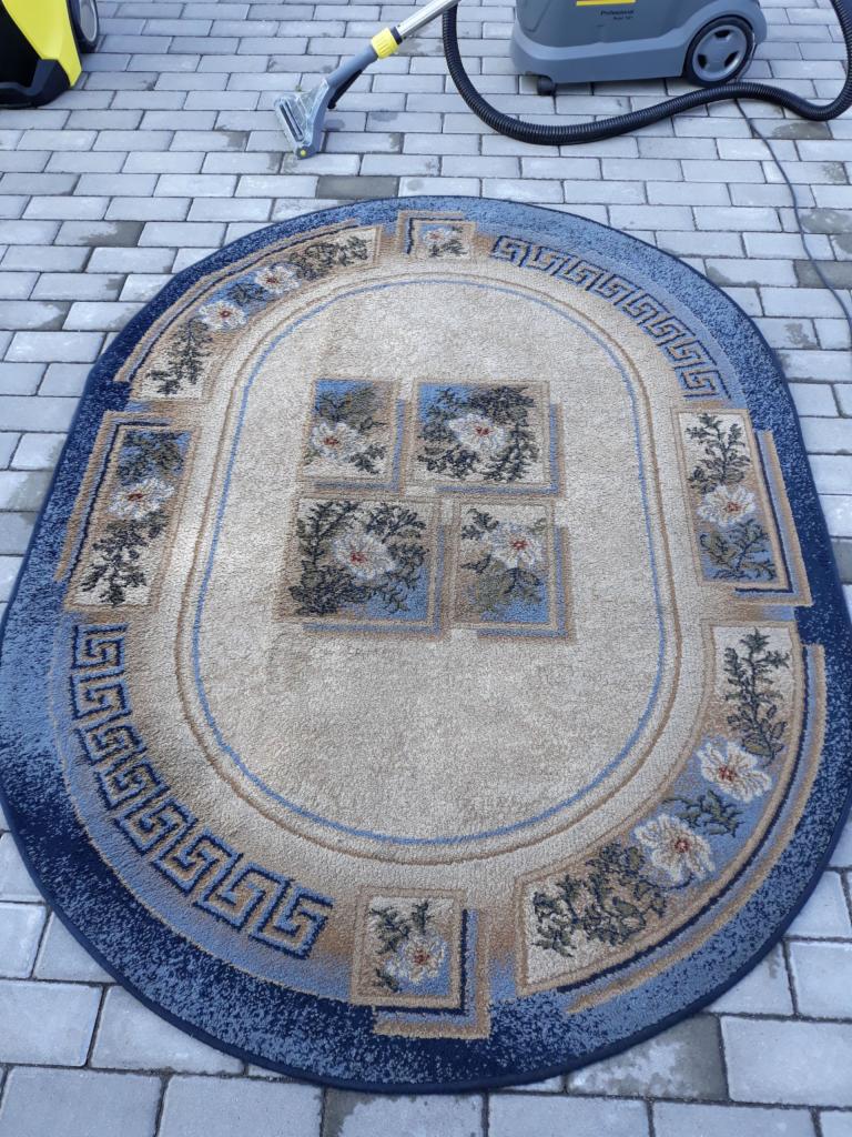 Owalny dywan z niebieskimi i beżowymi wzorami, leżący na szarej kostce brukowej, obok odkurzacza piorącego Karcher z końcówką do czyszczenia dywanów.