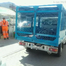 Niebieska ciężarówka z metalową klatką na pace, wypełniona odpadami, zaparkowana na chodniku z kostki brukowej. W tle pracownik w pomarańczowym kombinezonie wchodzi do zadaszonej konstrukcji.