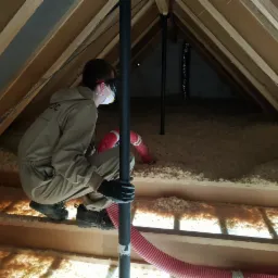 Pracownik w kombinezonie i masce ochronnej, w trakcie aplikacji izolacji włóknistej na poddaszu, widoczny czerwony wąż podający materiał izolacyjny.