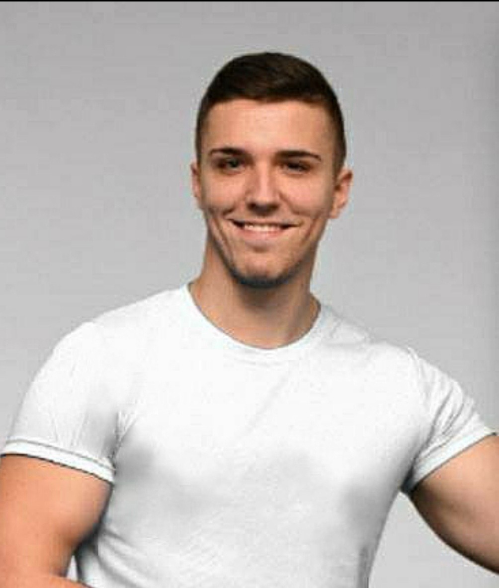 Uśmiechnięty mężczyzna w białym t-shircie na jasnoszarym tle, prezentujący sylwetkę.