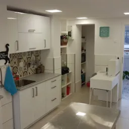 Wnętrze gabinetu weterynaryjnego z białymi meblami, stalowym stołem zabiegowym na pierwszym planie, dekoracyjną tabliczką z kotem i stetoskopem na ścianie oraz widokiem na schody prowadzące...