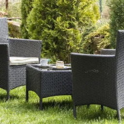 Zestaw mebli ogrodowych z plecionki technorattanowej: dwa fotele z poduszkami i stolik kawowy z filiżanką i ciastkiem, na trawniku w ogrodzie.