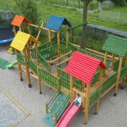 Kompleksowy plac zabaw z drewna i kolorowych dachów: zjeżdżalnie, mostki, huśtawki i trampolina w tle, dziecko na zjeżdżalni.