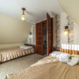 Pokój z dwoma łóżkami pojedynczymi, drewnianymi ramami, beżową pościelą i białymi poduszkami; na jednym łóżku leży żółty kwiat. Otwarta, ciemna drewniana stolarka drzwiowa prowadzi do kolejnego...