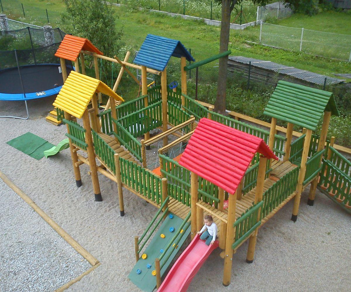 Kompleksowy plac zabaw z drewna i kolorowych dachów: zjeżdżalnie, mostki, huśtawki i trampolina w tle, dziecko na zjeżdżalni.