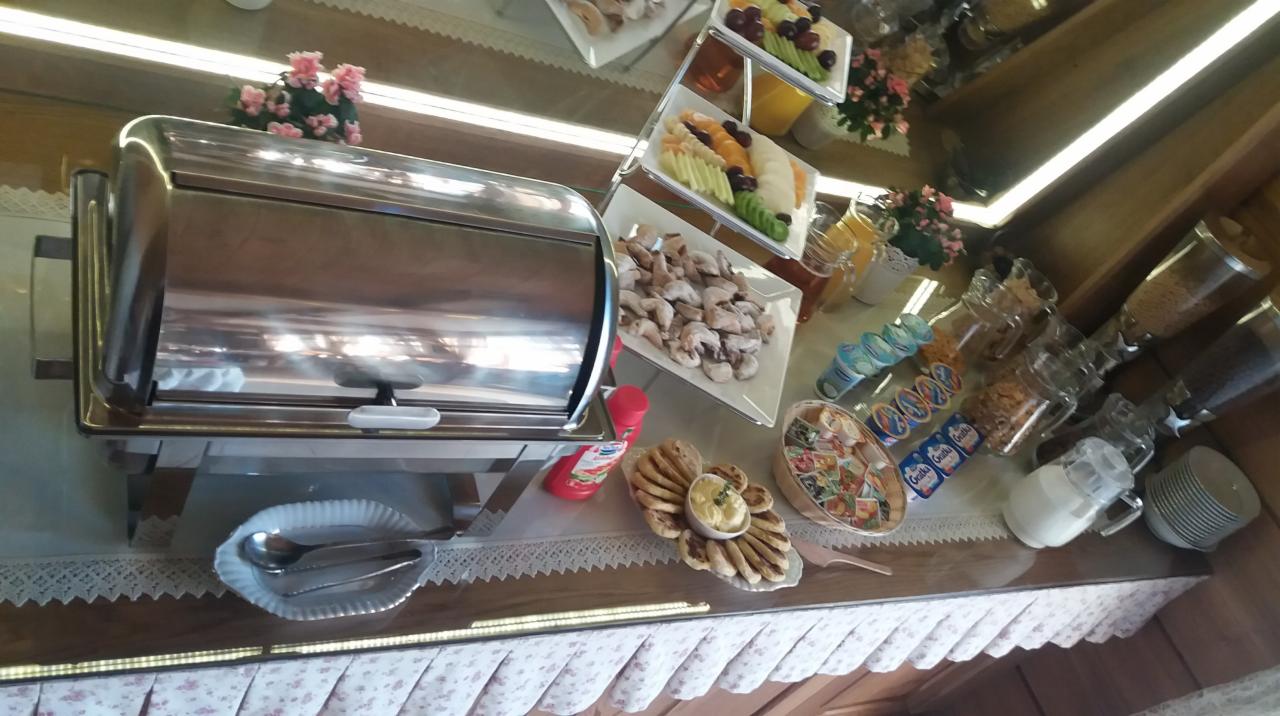 Szwedzki stół z metalowym podgrzewaczem, paterami z owocami, talerzami z przekąskami i pojemnikami na płatki śniadaniowe oraz jogurty, całość na drewnianym blacie z ozdobną falbanką.