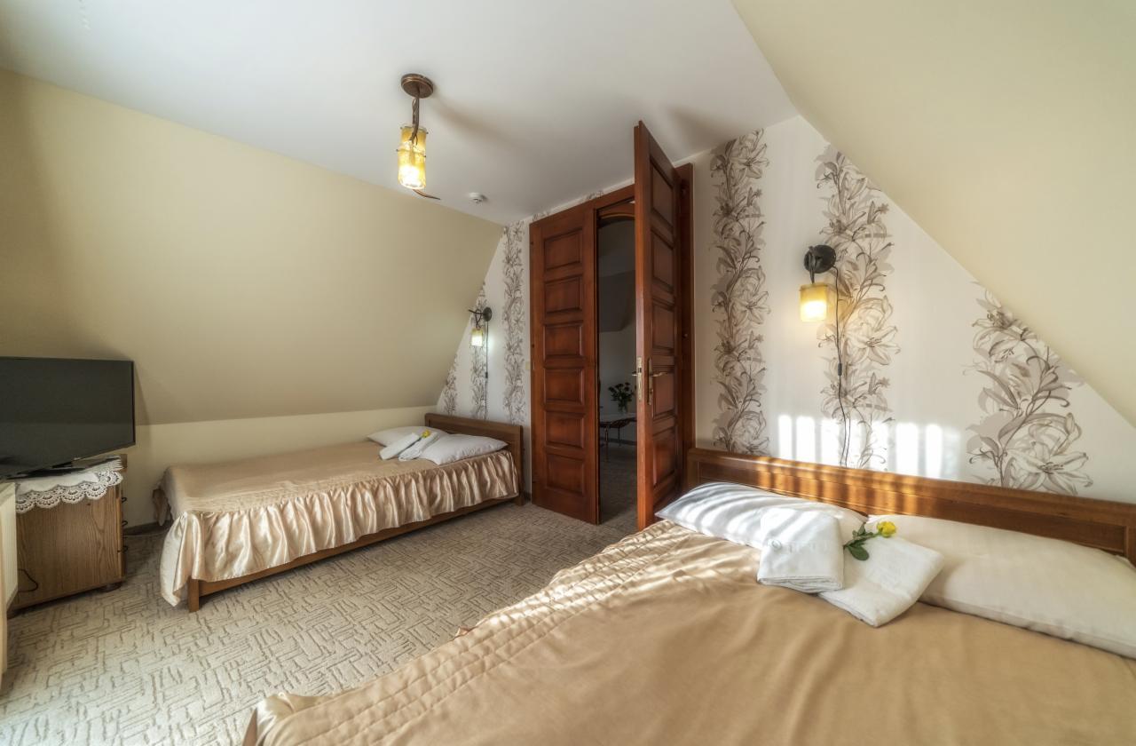 Pokój z dwoma łóżkami pojedynczymi, drewnianymi ramami, beżową pościelą i białymi poduszkami; na jednym łóżku leży żółty kwiat. Otwarta, ciemna drewniana stolarka drzwiowa prowadzi do kolejnego...