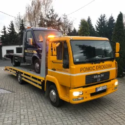 Żółta laweta MAN z niebieskim busem na platformie, zaparkowana na kostce brukowej przed budynkiem i zielonymi drzewami.