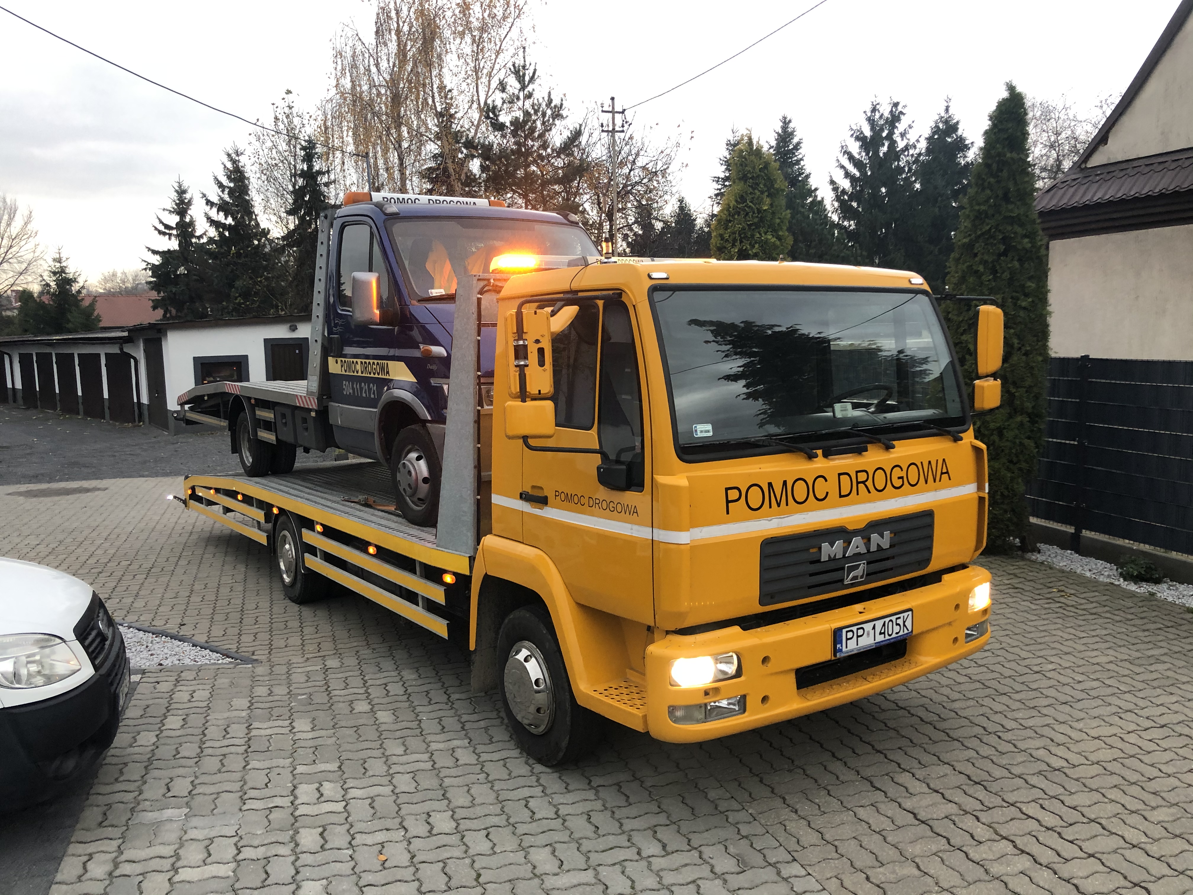 Żółta laweta MAN z niebieskim busem na platformie, zaparkowana na kostce brukowej przed budynkiem i zielonymi drzewami.