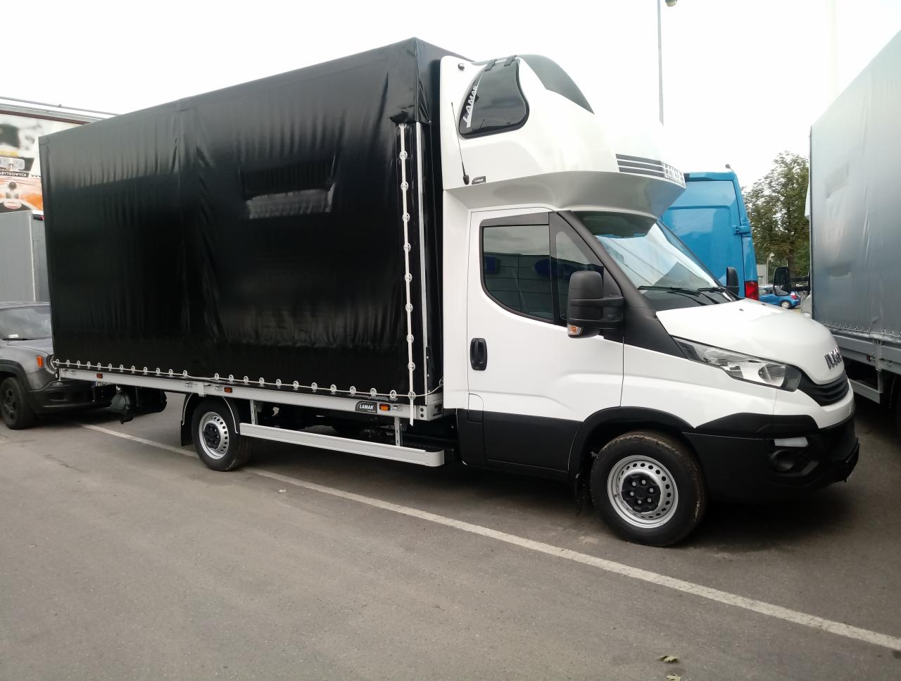 Biała ciężarówka z czarną plandeką na parkingu, widoczna również szara osobówka z tyłu, oświetlenie dzienne, lekko pochmurne niebo.