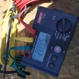 Miernik UNIT-T UT522 do pomiaru rezystancji uziemienia z podłączonymi przewodami pomiarowymi w kolorach czerwonym, żółtym i niebieskim, leżący na piaszczystym podłożu.