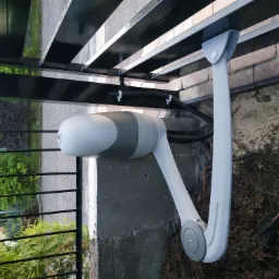 Zamontowany napęd ramieniowy do bramy skrzydłowej, widoczne okablowanie w peszlu przymocowane do ramy bramy, w tle zieleń ogrodu.