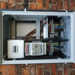 Otwarta, biała skrzynka elektryczna zamontowana na ceglanej ścianie, widoczne kable, bezpieczniki i inne komponenty elektryczne wewnątrz.