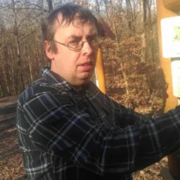 Mężczyzna w okularach i flanelowej koszuli stoi przy drewnianym słupie informacyjnym w lesie, jesienne liście na ziemi, widoczne drzewa bez liści w tle.