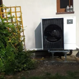 Biała jednostka zewnętrzna z czarnym wentylatorem, zamontowana na metalowej konstrukcji przy ścianie domu z drewnianymi oknami i kratownicą porośniętą roślinami.