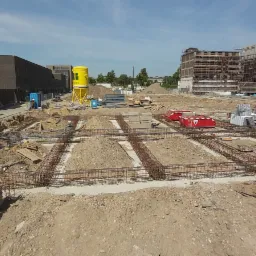 Widok z góry na plac budowy z przygotowanymi fundamentami, stalowymi zbrojeniami, żółtym zbiornikiem na wodę i budynkami w tle.