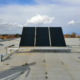 Trzy panele słoneczne zamontowane na płaskim dachu z widocznymi rurami i odpowietrznikami, w tle budynki i drzewa pod błękitnym niebem z chmurami.