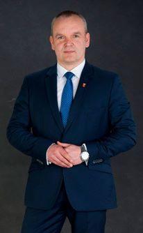 Portret mężczyzny w granatowym garniturze i niebieskim krawacie, z zegarkiem na lewym nadgarstku i przypinką na klapie, na ciemnoszarym tle.