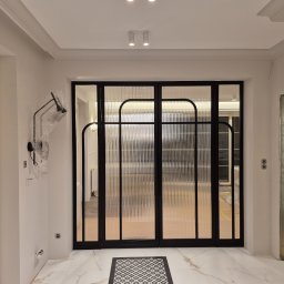 BB Fabrik - Nowoczesne, czteroskrzydłowe drzwi wewnętrzne z ryflowanego szkła i czarnych profili, na marmurowej posadzce z dywanikiem. Widok na sąsiednie pomieszczenia.