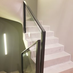 BB Fabrik - Minimalistyczne schody z jasnymi stopniami i szklaną balustradą z ciemną ramą. Nowoczesne oświetlenie ścienne. Ujęcie z dołu, podkreślające geometrię.