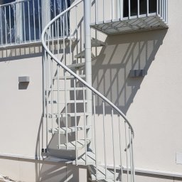 BB Fabrik - Zewnętrzne, metalowe schody spiralne w jasnym odcieniu szarości, prowadzące na balkon z balustradą. Widoczne detale konstrukcyjne i cień rzucany na elewację budynku.