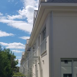 BB Fabrik - Białe schody spiralne na zewnątrz budynku z jasną elewacją i szarym dachem, widok z dołu na tle błękitnego nieba z chmurami.