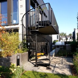 BB Fabrik - Zewnętrzne, spiralne schody metalowe w kolorze czarnym, prowadzące na balkon nowoczesnego domu z białej cegły. Widoczne elementy zieleni i kostka brukowa.