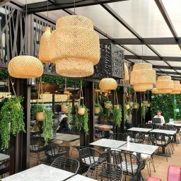 BB Fabrik - Zadaszony taras kawiarni z rattanowymi lampami i zielonymi roślinami. Metalowa konstrukcja dachu i ażurowe panele dekoracyjne. W tle osoba pracująca na laptopie.