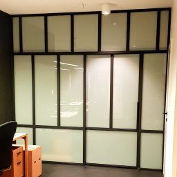 BB Fabrik - Przeszklona ścianka działowa z czarnymi profilami w biurze. Matowe szkło zapewnia prywatność. Widoczny fragment biurka i krzesła.