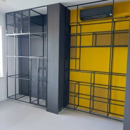 BB Fabrik - Czarne, metalowe regały z siatką na żółtej ścianie, nowoczesny design. Minimalistyczne półki z metalu w industrialnym stylu, widok z dołu, jasne wnętrze.