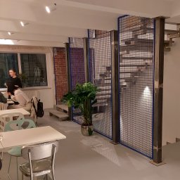 BB Fabrik - Industrialne schody z metalową siatką w kawiarni. Stalowe słupy, niebieska siatka, betonowe stopnie. W tle osoby przy stolikach i okno.