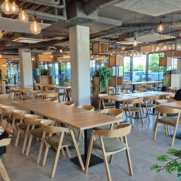 BB Fabrik - Nowoczesne wnętrze restauracji z drewnianymi stołami i krzesłami, oświetlone wiszącymi żarówkami. Widoczne instalacje pod sufitem i duże okna.