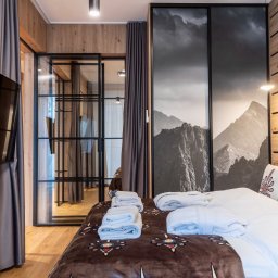 BB Fabrik - Nowoczesna sypialnia z drewnianą ścianą i czarną, przeszkloną ścianką działową. Widoczne łóżko z góralskim wzorem i dekoracyjna grafika gór.