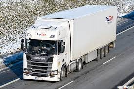 Biała ciężarówka marki Scania z naczepą, z logo firmy TopTen, jadąca po asfalcie w zimowej scenerii. Widoczny kierowca za kierownicą.